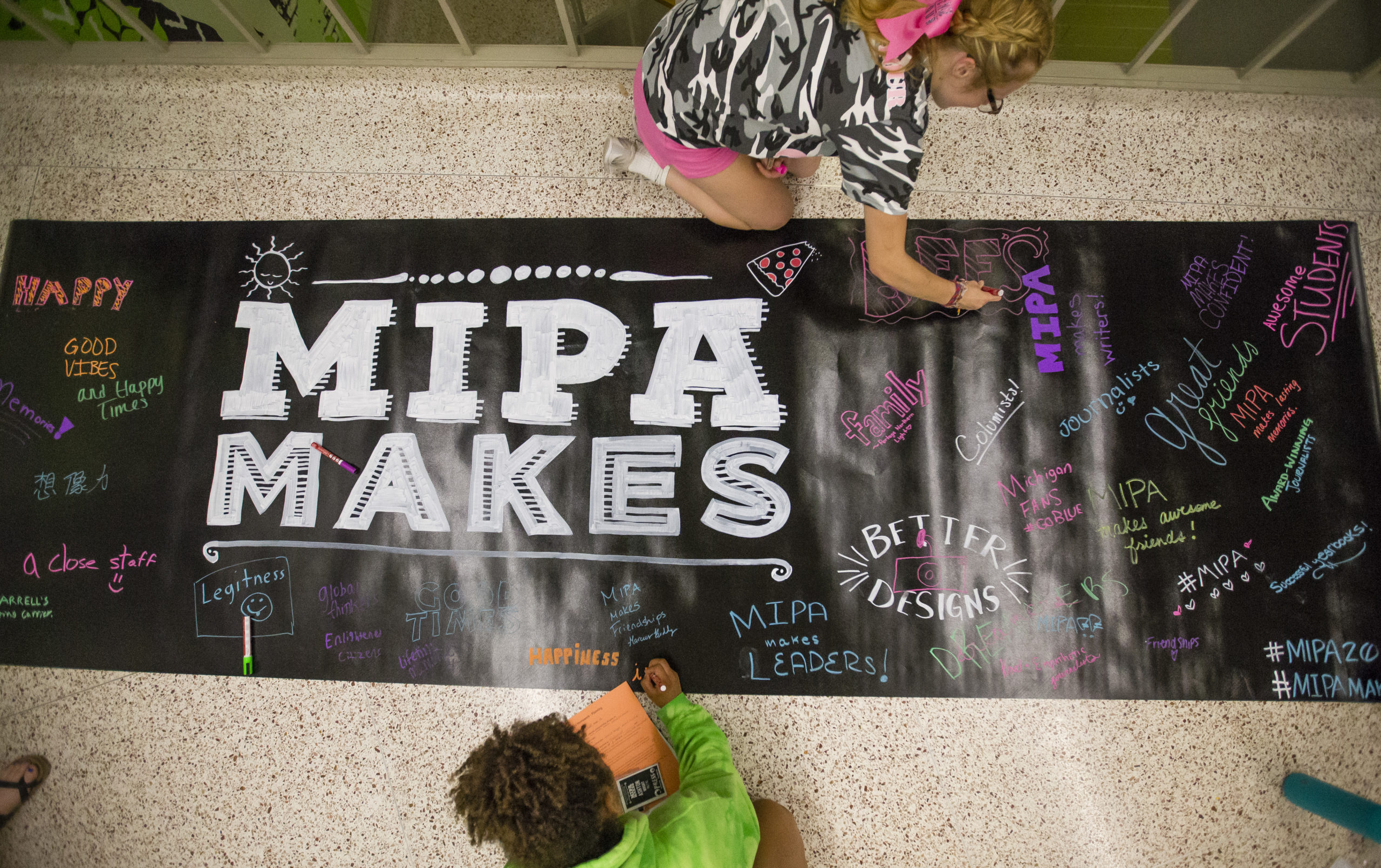 Two young people work on a banner on the floor that reads "MIPA Makes"